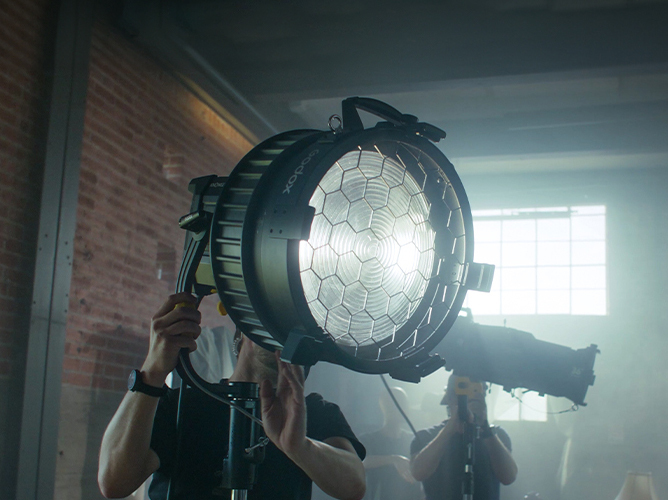 To personer justerer en stor studiobelysning med bikubemønster foran, i et rom med murvegger og vinduer som slipper inn mykt lys