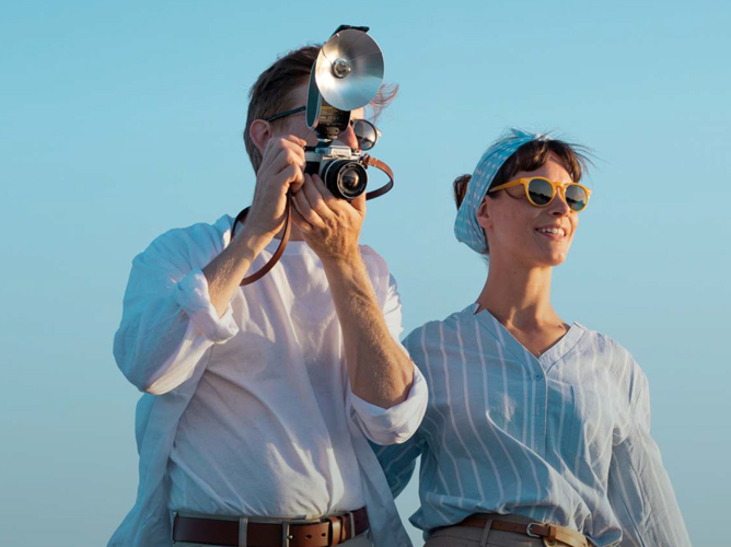En mann i hvit skjorte tar et bilde med et vintagekamera mens han står ved siden av en kvinne i lyseblå stripete skjorte under en blå himmel