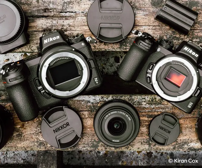 To Nikon speilløse kameraer med tilbehør som objektivdeksler, batteri og adapter plassert på en rustikk trebordsoverflate