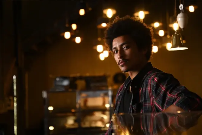 En person med mørkt krøllete hår står inne i et moderne rom opplyst av varme lyspærer, med bakgrunn som viser hyller og en koselig atmosfære