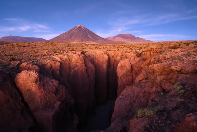En dyp kløft i rødt ørkenlandskap med en vulkan i bakgrunnen under en klar blå himmel