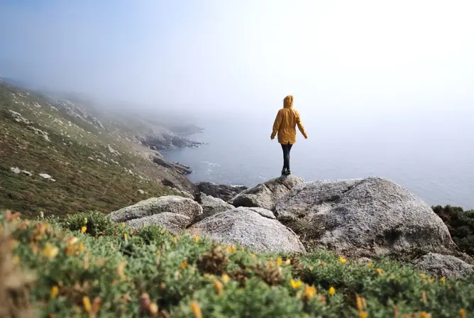 En person i gul jakke står på en stein med utsikt mot havet og tåkelagte fjell. En person i gul jakke står på en stein med utsikt mot havet og tåkelagte fjell.