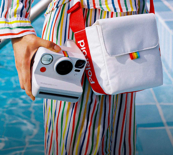 Person i fargerik stripete drakt holder et hvitt Polaroid kamera og matchende veske med regnbuedetaljer nær et basseng