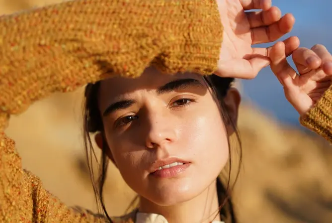 Person med brun strikkegenser holder hendene opp i sollyset med en gyllen bakgrunn.