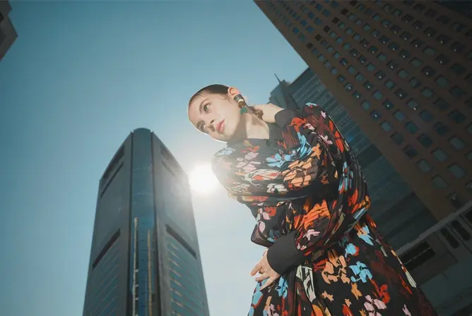 En person i fargerike klær poserer foran høye moderne bygninger med solen i bakgrunnen
