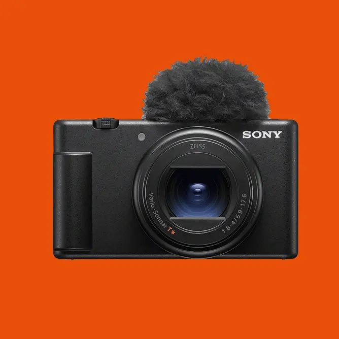 Svart Sony-kamera med en stor linse og en mikrofon med pelstrekk på oransje bakgrunn