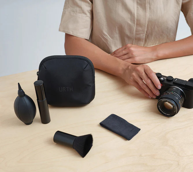 Person bruker et kamera på et bord med et svart rensekit, inkludert børste, klut og luftblåser Person bruker et kamera på et bord med et svart rensekit, inkludert børste, klut og luftblåser