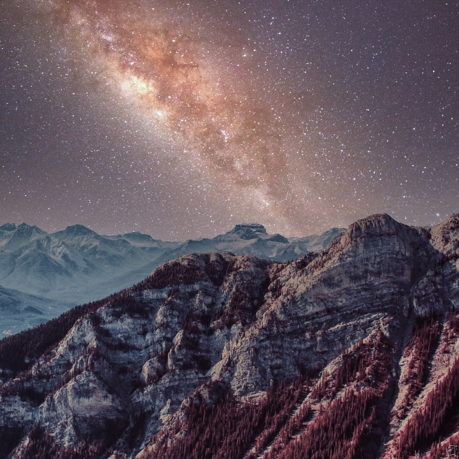 En nattehimmel med melkeveien som lyser klart, over fjelltopper og mørke furutrær