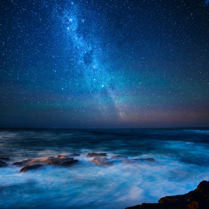 Stjerneklar nattehimmel over et rolig hav med glitrende bølger som slår mot steiner
