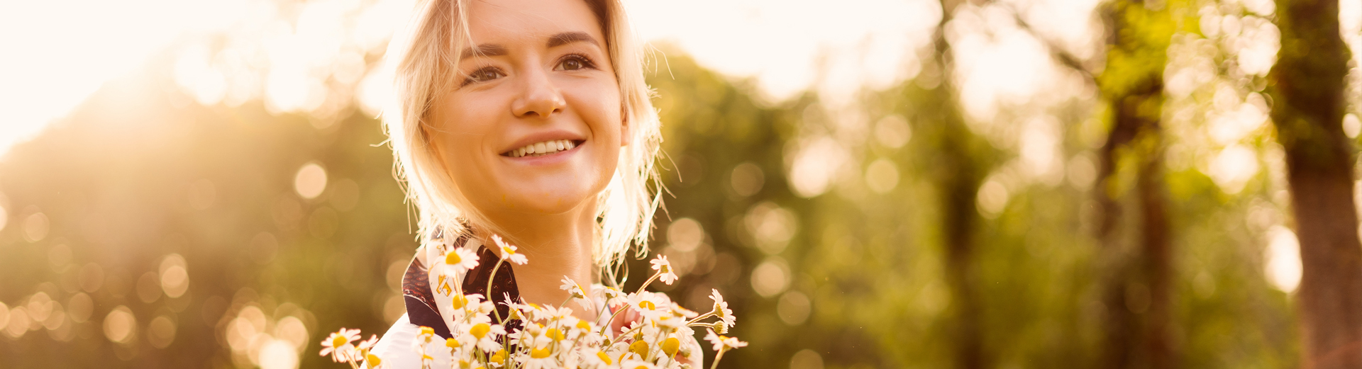 Person med blondt hår holder en bukett med prestekrager i solnedgang i skogen