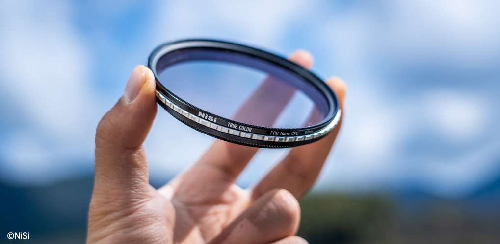 Hånd holder et kamera-filter mot en blå himmel med hvite skyer i bakgrunnen