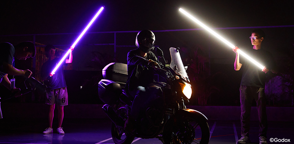 En motorsyklist med hjelm sitter på en sykkel i mørket, med lilla og hvite lysrør holdt av to personer i bakgrunnen En motorsyklist med hjelm sitter på en sykkel i mørket, med lilla og hvite lysrør holdt av to personer i bakgrunnen