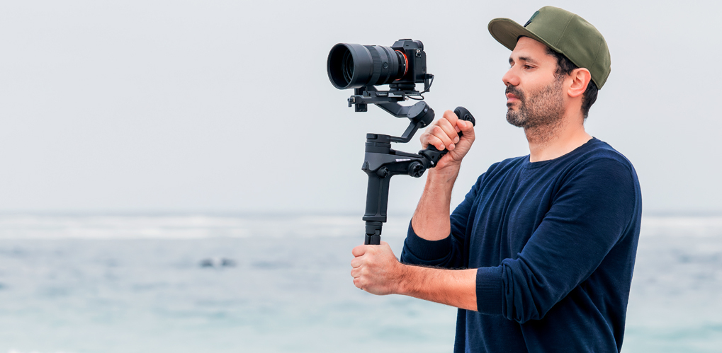 En person holder et kamera med en stabilisator foran et hav i bakgrunnen