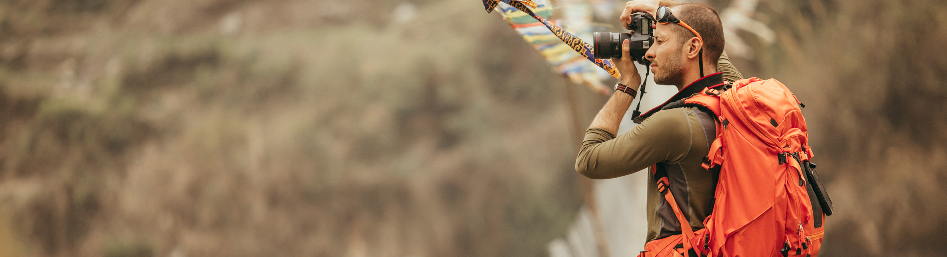 En person tar bilder med et speilreflekskamera, iført en oransje tursekk i et naturlandskap med fargerike vimpler i bakgrunnen