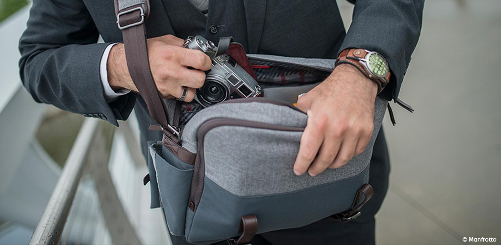 En mann i dress tar et kamera ut av en grå skulderveske med lærdetaljer