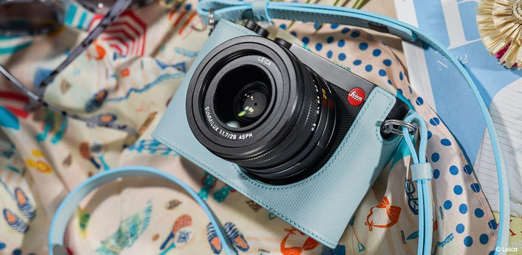 Lyseblå Leica-kamera med matchende kamerastropp ligger på et fargerikt, mønstret stoff.