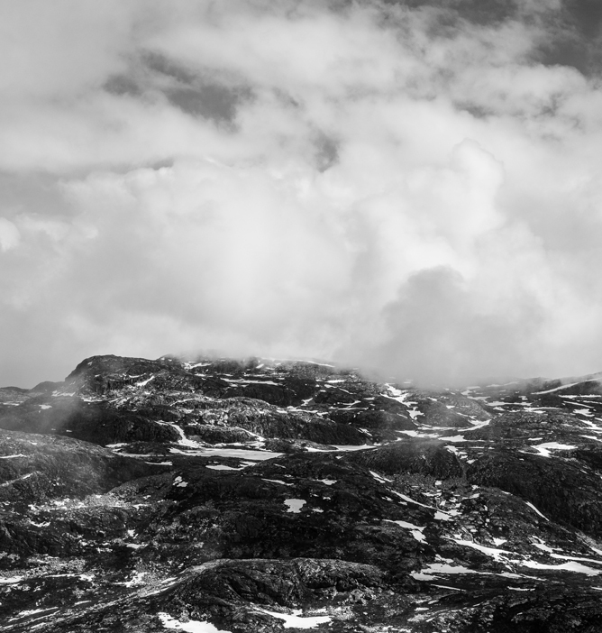 Svarthvitt fjellandskap med steinete overflater og lette snøflekker under en dramatisk himmel med skyer