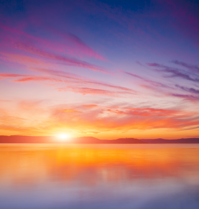Solnedgang med myke rosa, oransje og lilla skyer over en stille innsjø