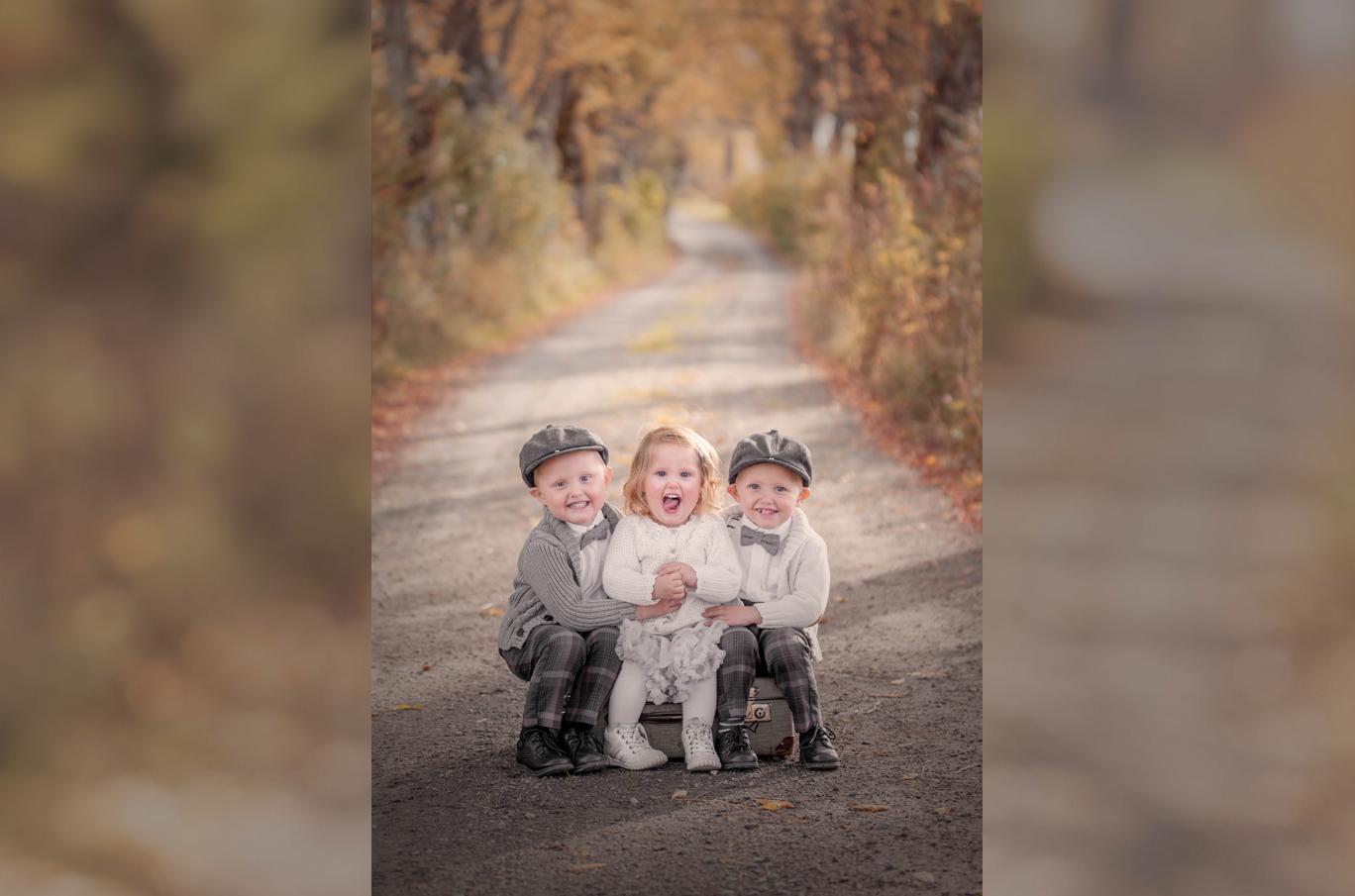 Tre barn sitter sammen på en gammel koffert midt på en grusvei omringet av høstfarger.