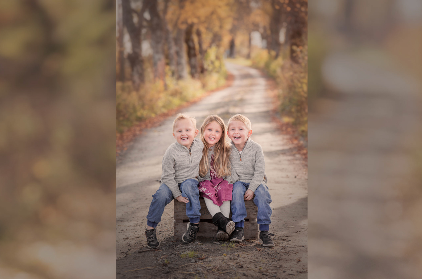 Tre barn i solfylt høstlandskap, sittende sammen på en trekasse midt på en grusvei omringet av gule og oransje høstblader