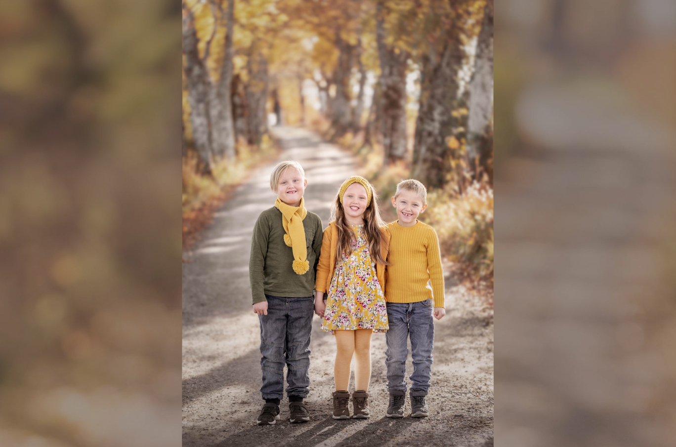 Tre barn står på en grusvei omgitt av høstfargede trær; alle har på seg klær i varme gule og grønne toner.