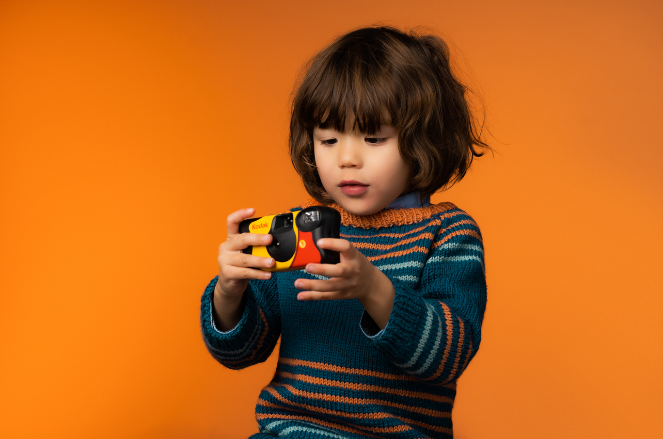 Barn i stripete genser holder et fargerikt Kodak-kamera med begge hender mot en oransje bakgrunn
