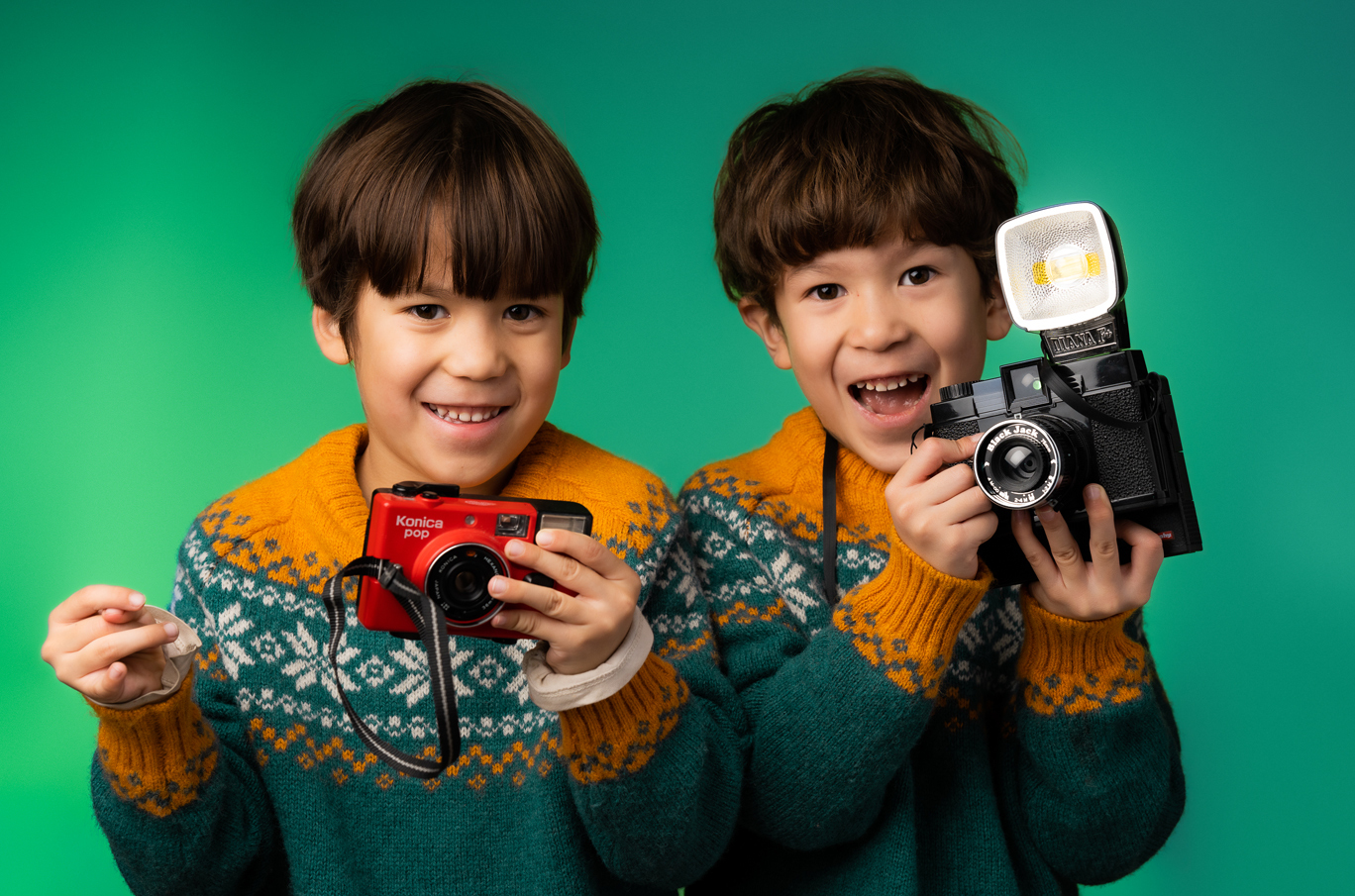 To barn i matchende gensere med mønster holder hver sin retro kamera foran en grønn bakgrunn