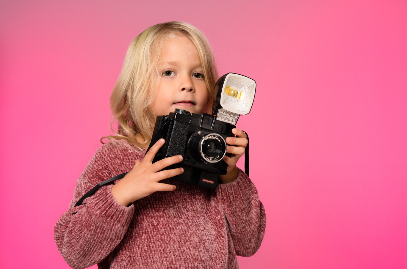 Et barn med langt blondt hår holder et klassisk kamera med stor blits mot en rosa bakgrunn.