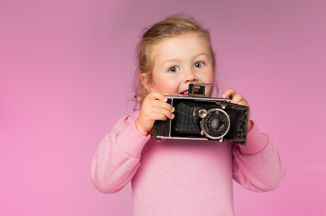 Liten jente i rosa genser holder et gammeldags kamera mot et rosa bakteppe