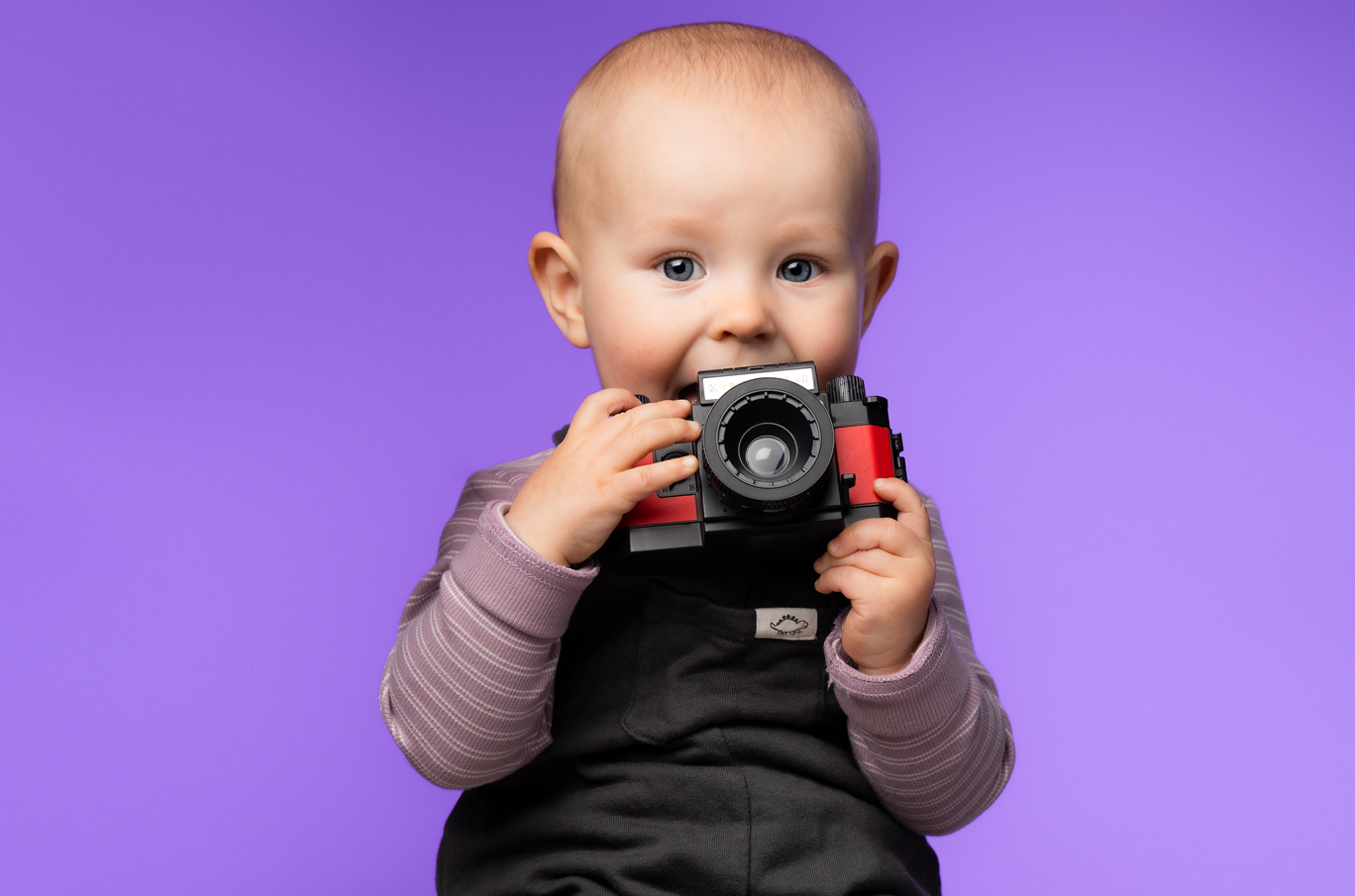 Baby i stripete genser holder et fargerikt lekelekamera mot en lilla bakgrunn
