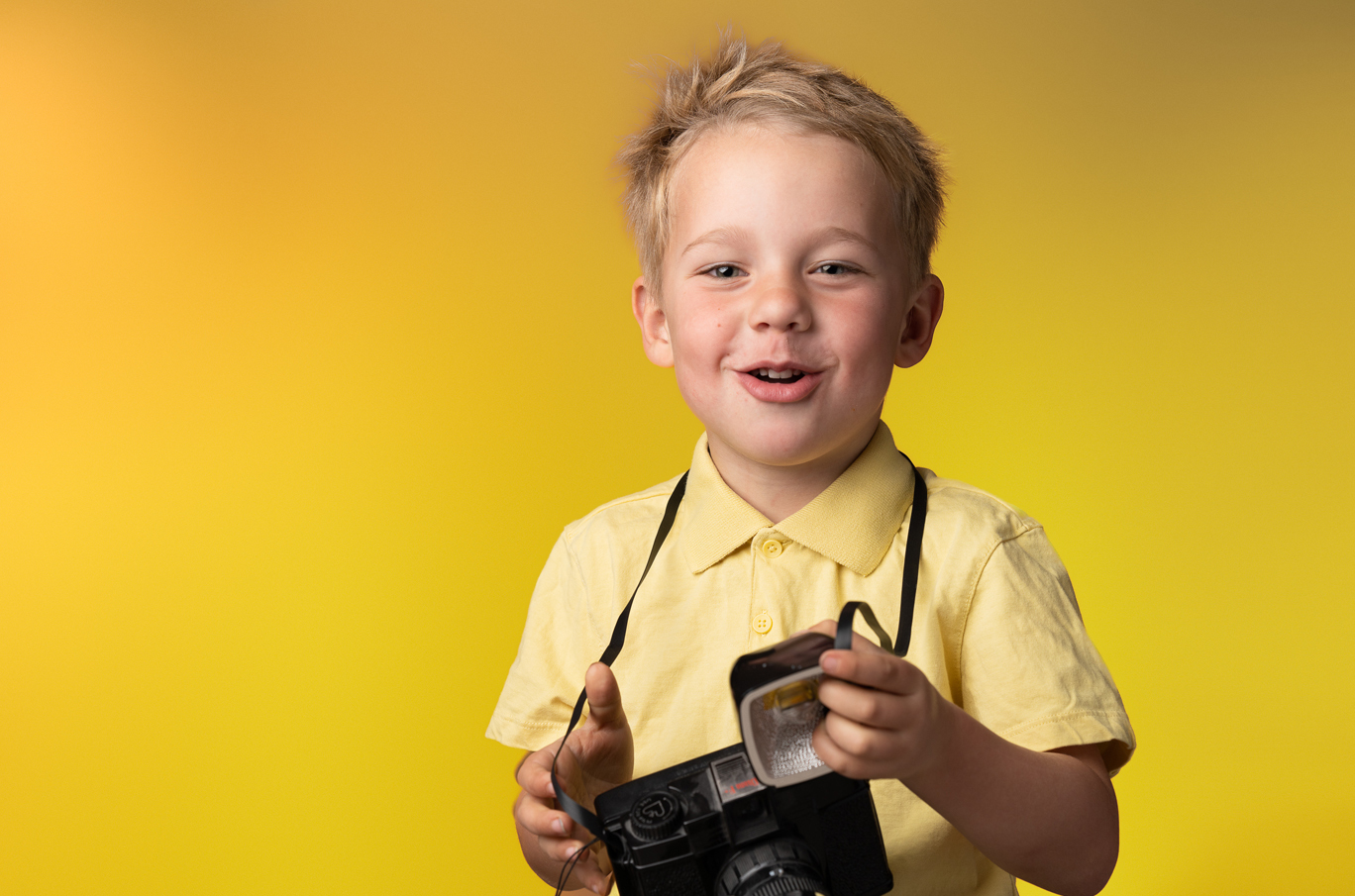 Liten gutt i gul skjorte holder et gammeldags kamera foran en gul bakgrunn