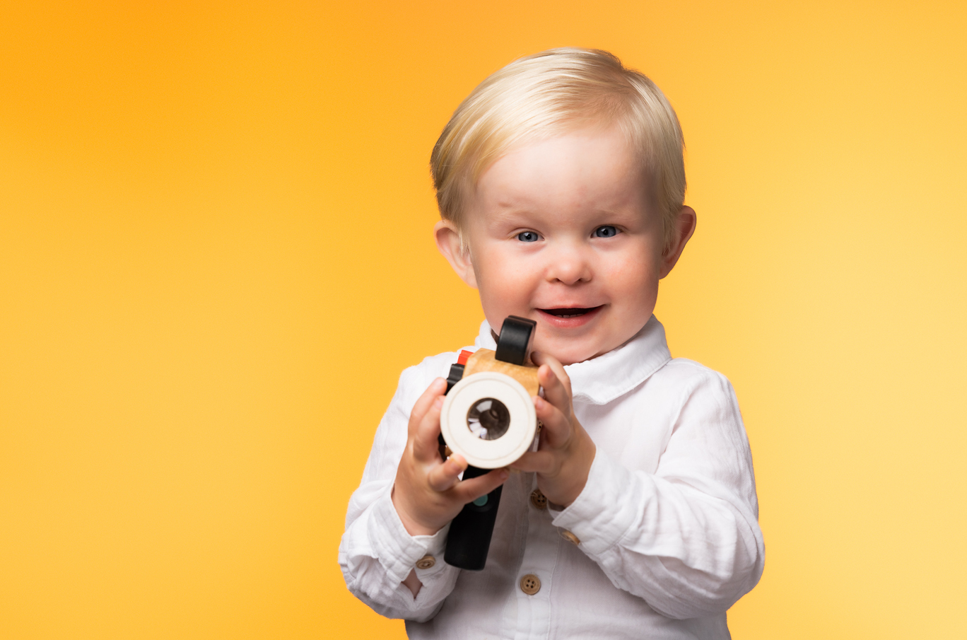 Et barn i hvit skjorte holder et treleketøykamera mot en varm, gul bakgrunn