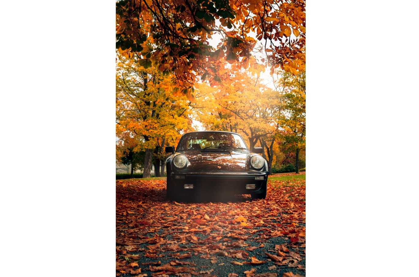 En brun klassisk bil står parkert på en vei dekket av oransje høstløv, omgitt av trær med gyldent løvverk