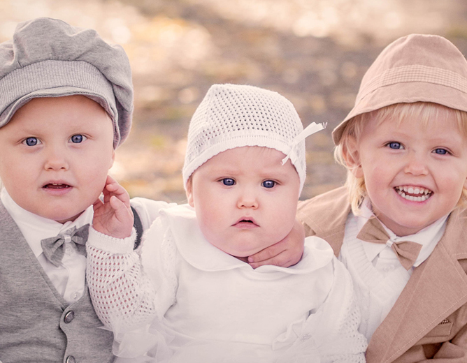 Tre små barn i vintage-inspirerte klær og hodeplagg sitter sammen utendørs på en solrik dag.