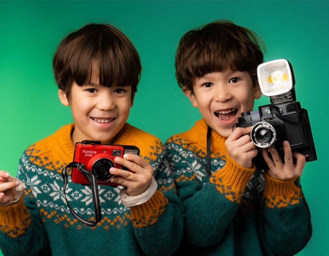 To barn i matchende gensere med mønster som holder hvert sitt kamera, én rød og én svart