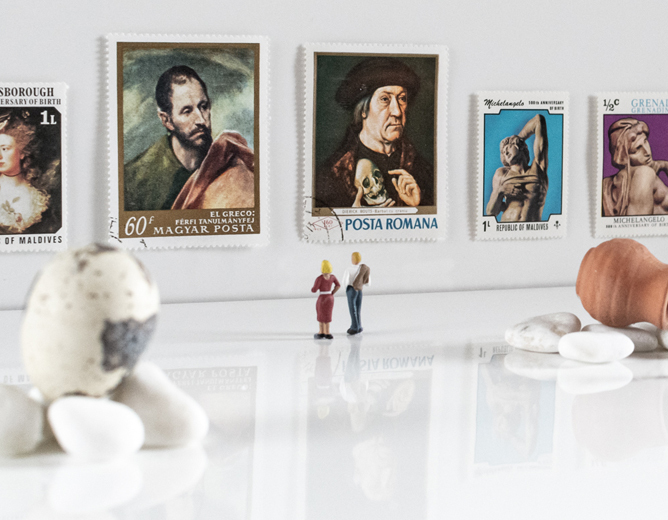 To frimerker med kunstverk i en bakgrunn, små miniatyrfigurer som står foran, og dekorative egg og steiner på en blank overflate