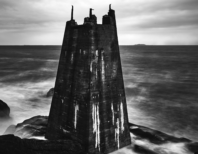Et massivt, gammelt fyrtårn av stein står på en klippe ved havet under en overskyet himmel