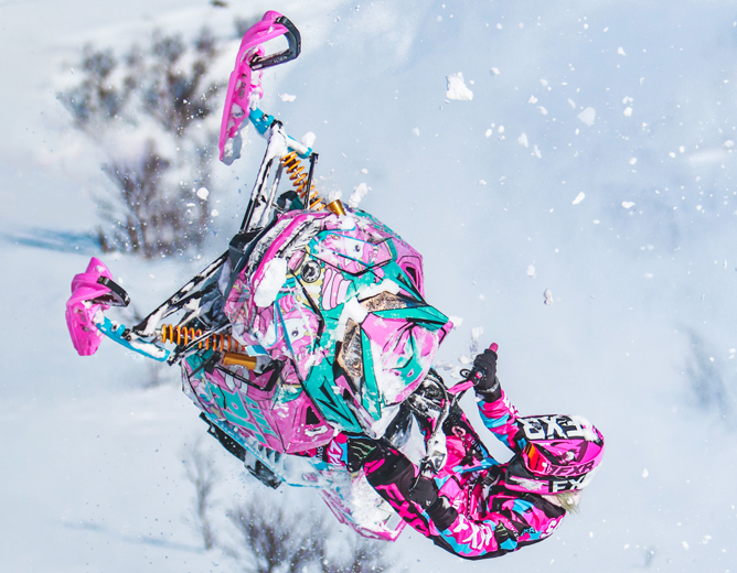 Person kjører snøscooter med fargerikt rosa og blått mønster, utfører et luftig hopp i snødekt terreng