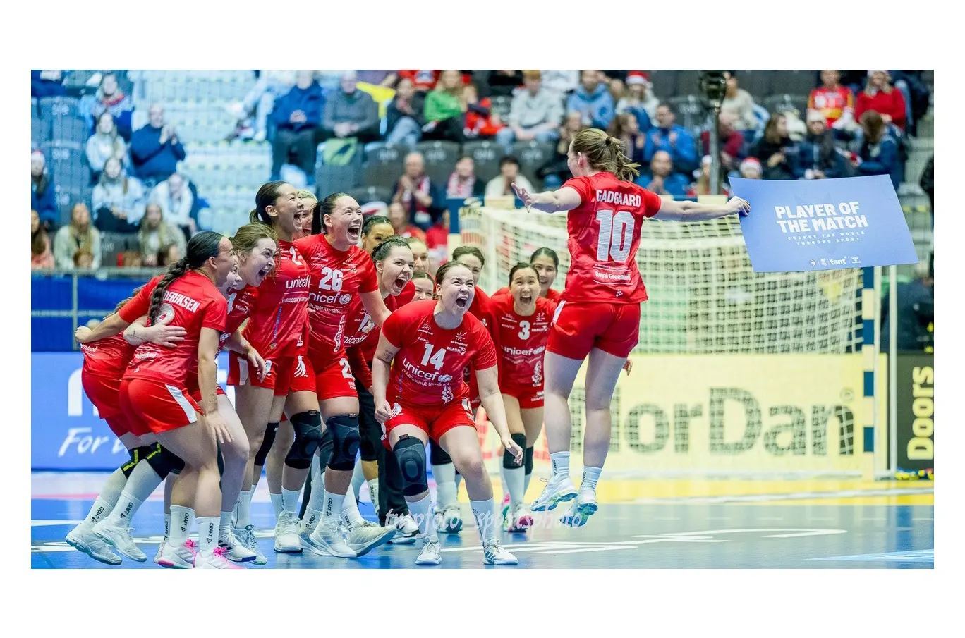 Håndballspillere i røde drakter feirer på banen, en spiller holder en "Player of the Match"-plakat
