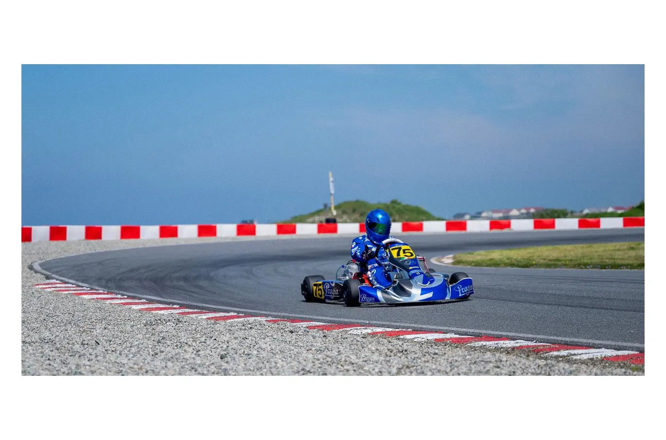 En gokartfører med blå hjelm og drakt kjører gjennom en skarp sving på en racerbane med rød-hvite barrierer