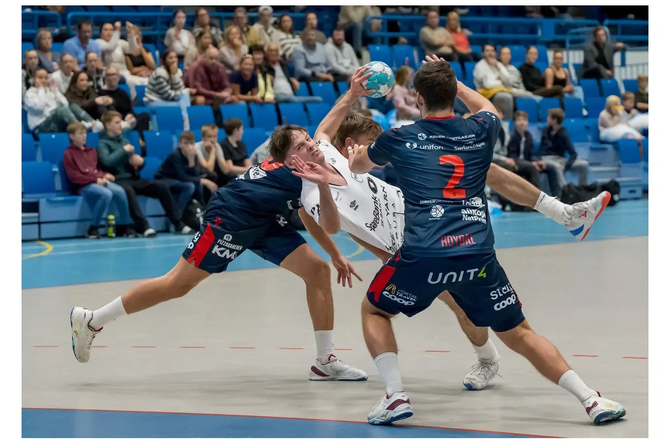 To håndballspillere i blått forsvarer mot en spiller i hvitt som holder en ball i en kamp på en innendørs arena