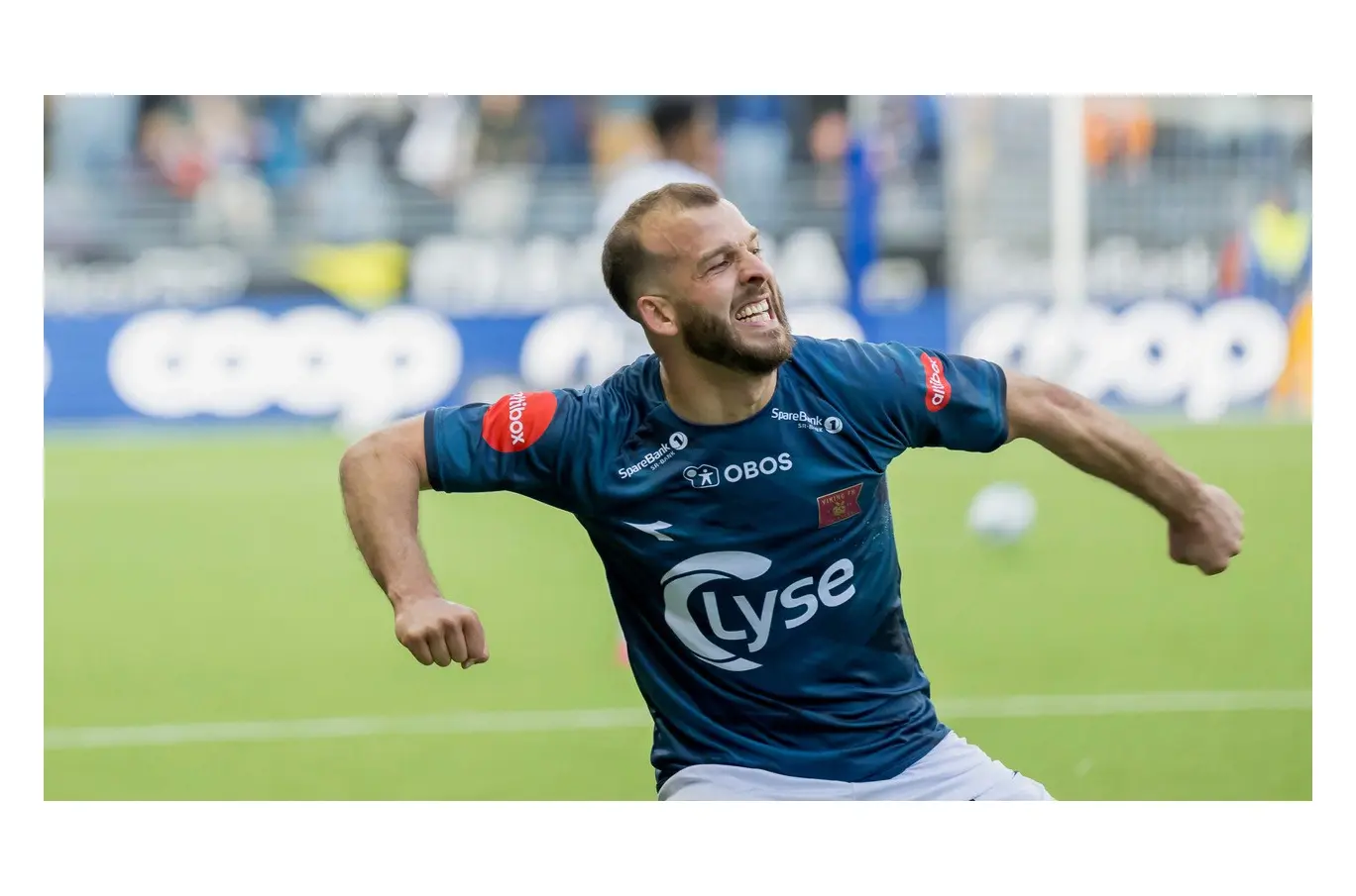 Fotballspiller i blå drakt jubler med utstrakte armer på gressbane.