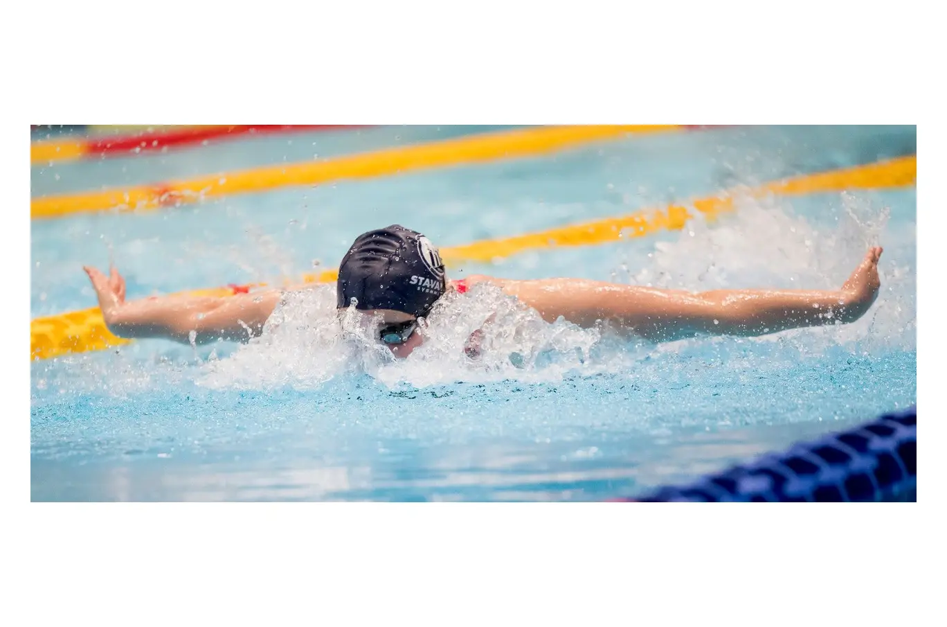 En svømmer i butterfly-stil midt i et basseng, iført mørk badehette og svømmebriller