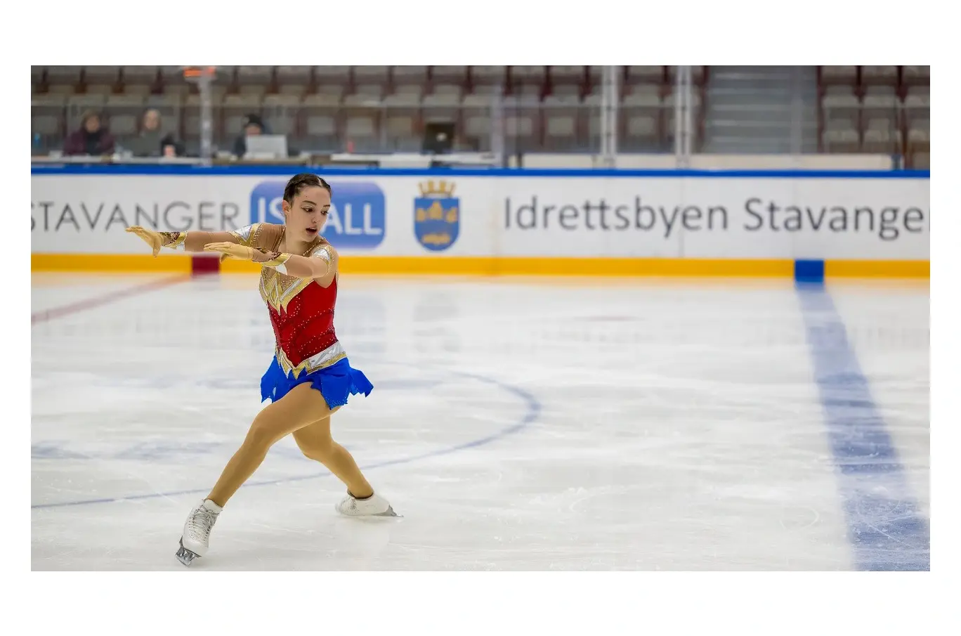 Kvinnelig kunstløper i fargerik rød og blå drakt utfører et elegant skøyteelement på isen