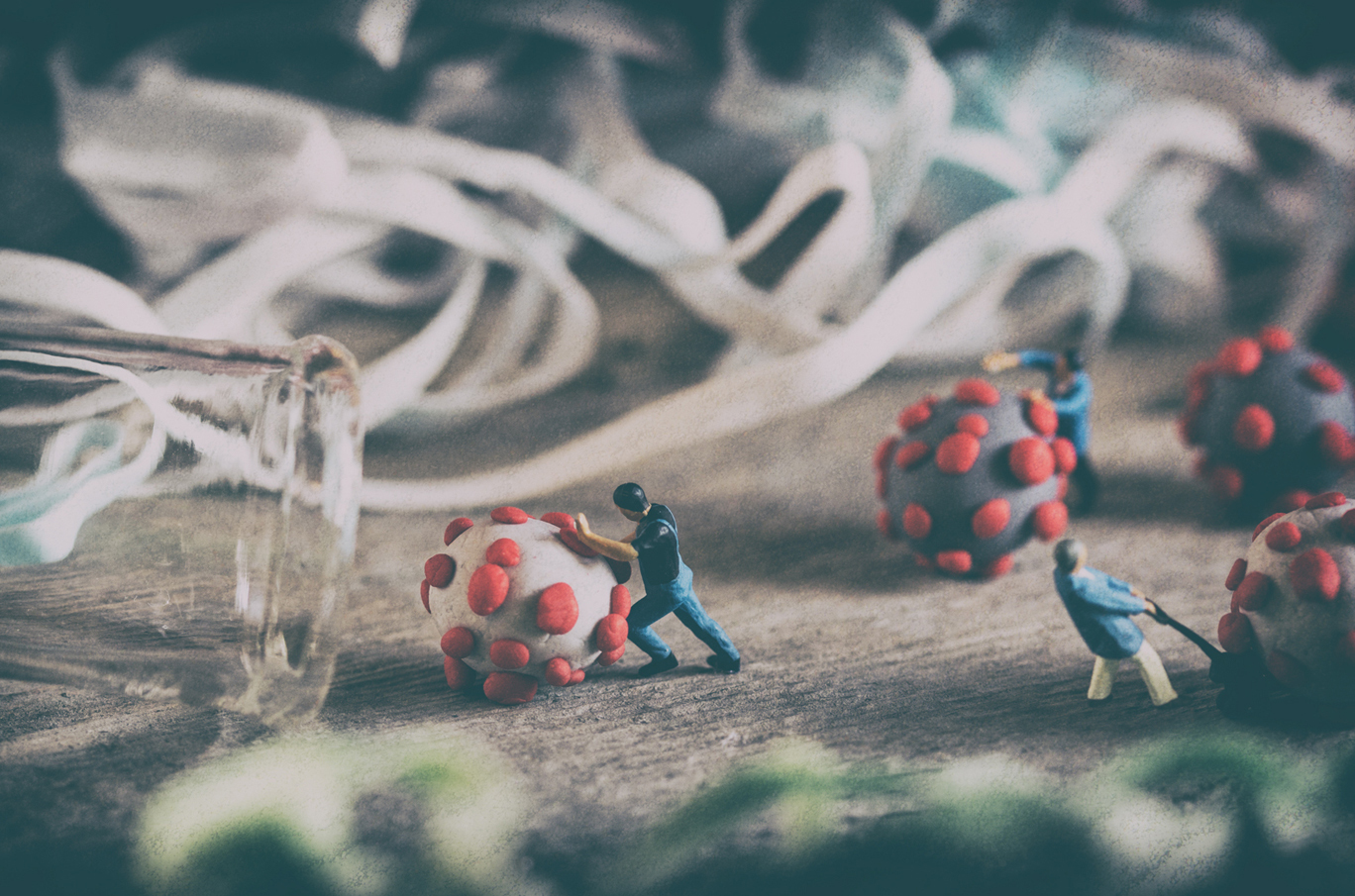 Minatyrmennesker skyver store, modellbaserte viruskuler med røde utvekster, omringet av hvite bånd og et veltet glass