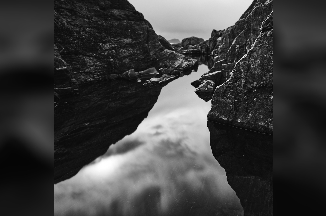 Svart-hvitt bilde av steinformasjoner som reflekteres i stille vann med en skyet himmel i bakgrunnen