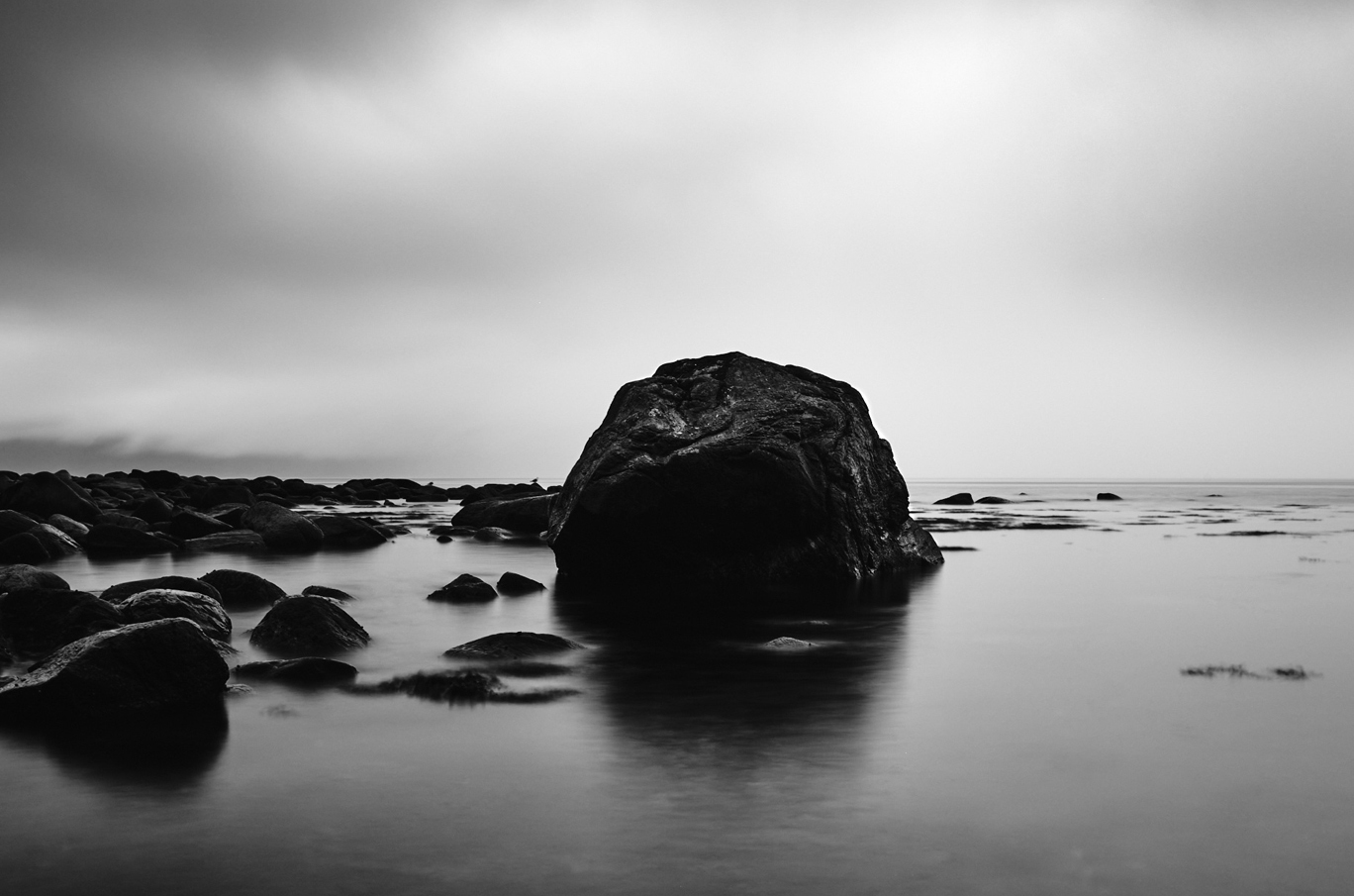 Stor stein omgitt av mindre steiner i et rolig hav, svart-hvitt landskap