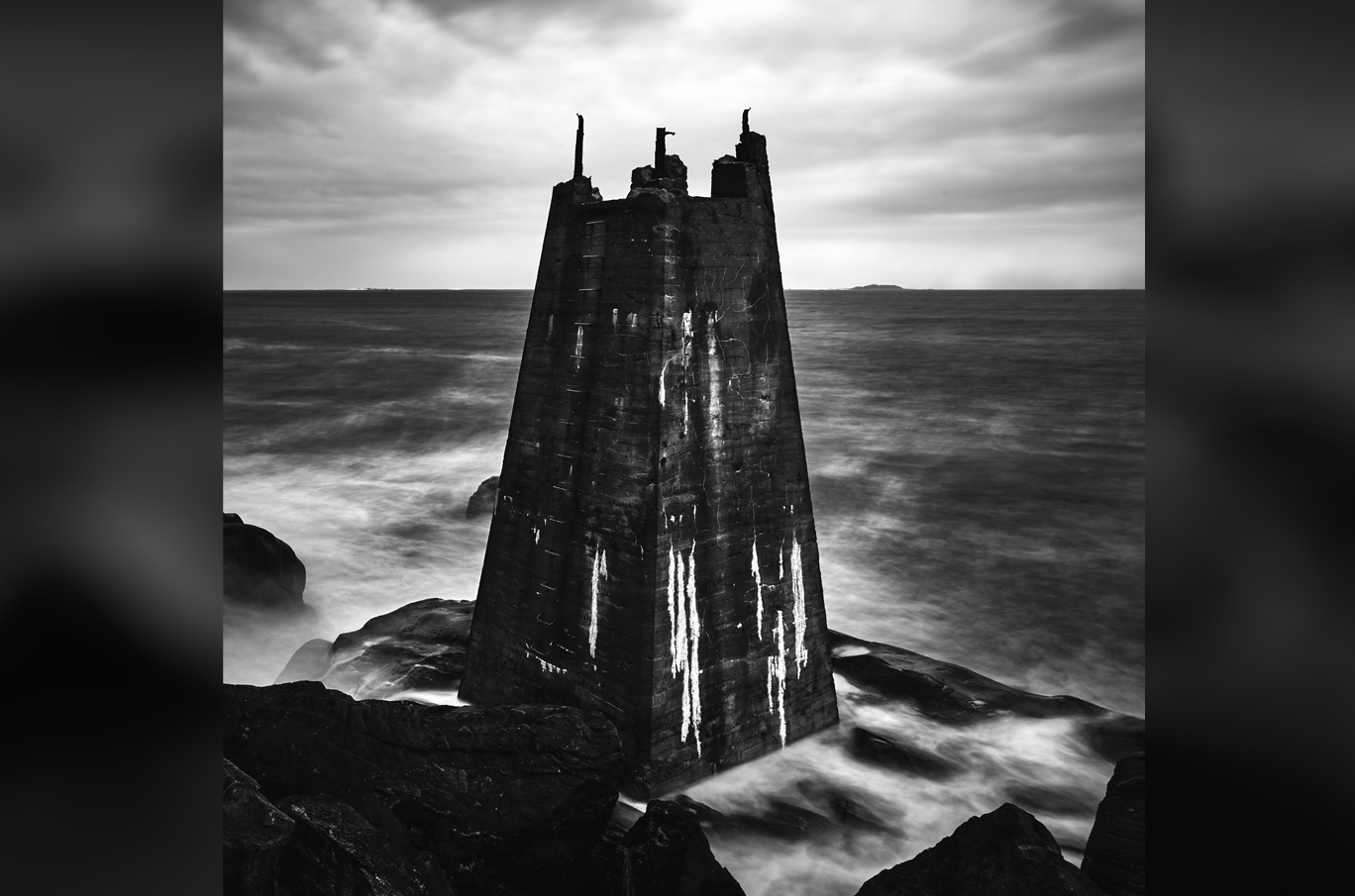 Svart-hvitt bilde av et robust, ødelagt tårn stående på klipper omgitt av bølgende hav