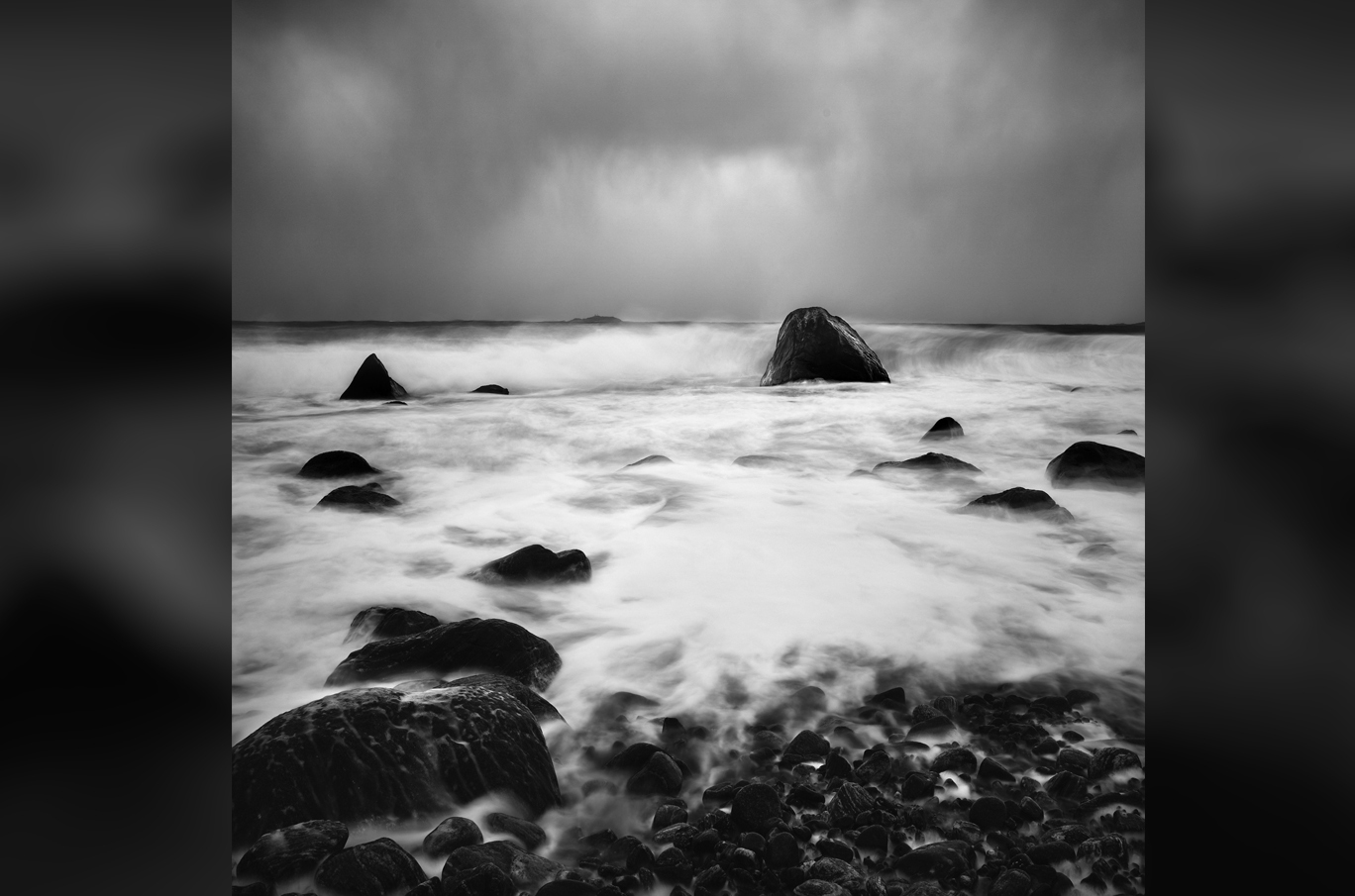 Svart-hvitt bilde av steiner på en strand med bølger som skyller over dem i tåkeaktig vær