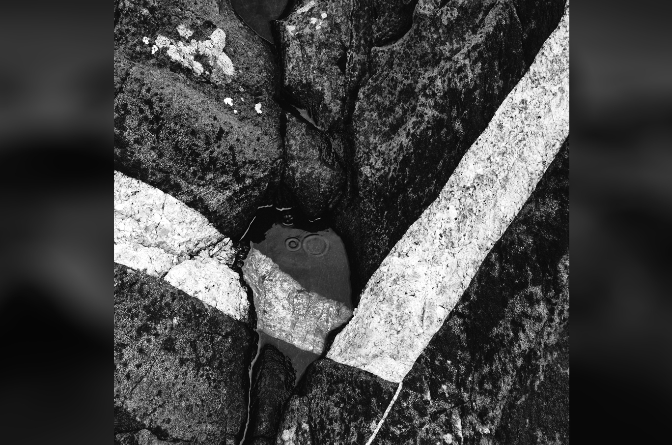 Svart-hvitt bilde av steiner med sprukne overflater og et lite vannspeil midt i kløften